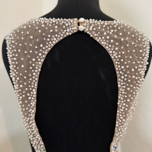 May include: A beige mesh fabric with a large open back design, embellished with white pearls and rhinestones. The fabric is attached to a black mannequin.