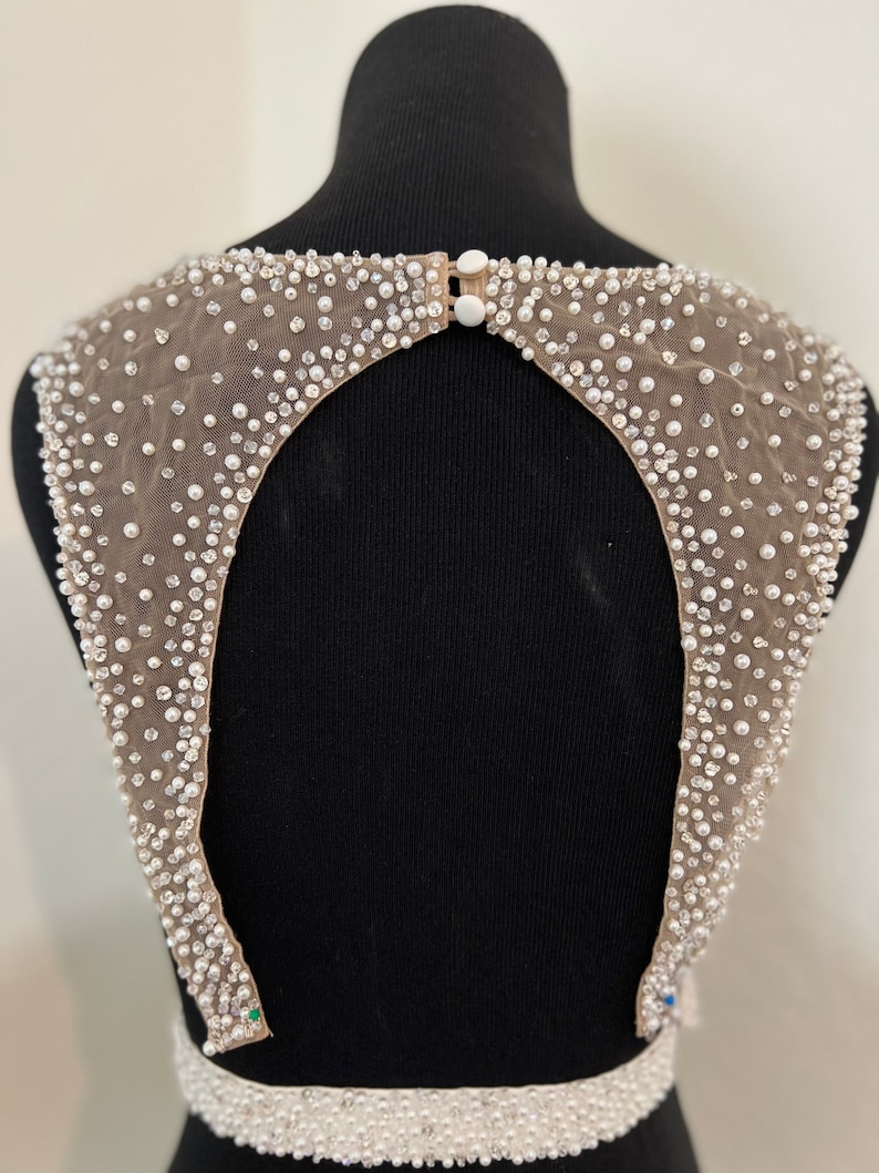 May include: A beige mesh fabric with a large open back design, embellished with white pearls and rhinestones. The fabric is attached to a black mannequin.