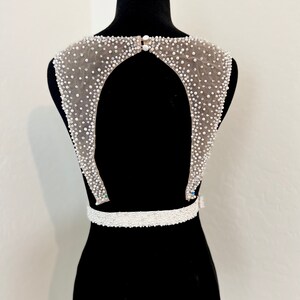 May include: A white beaded bridal belt with a low back design. The belt features a beaded neckline and a beaded waistband.
