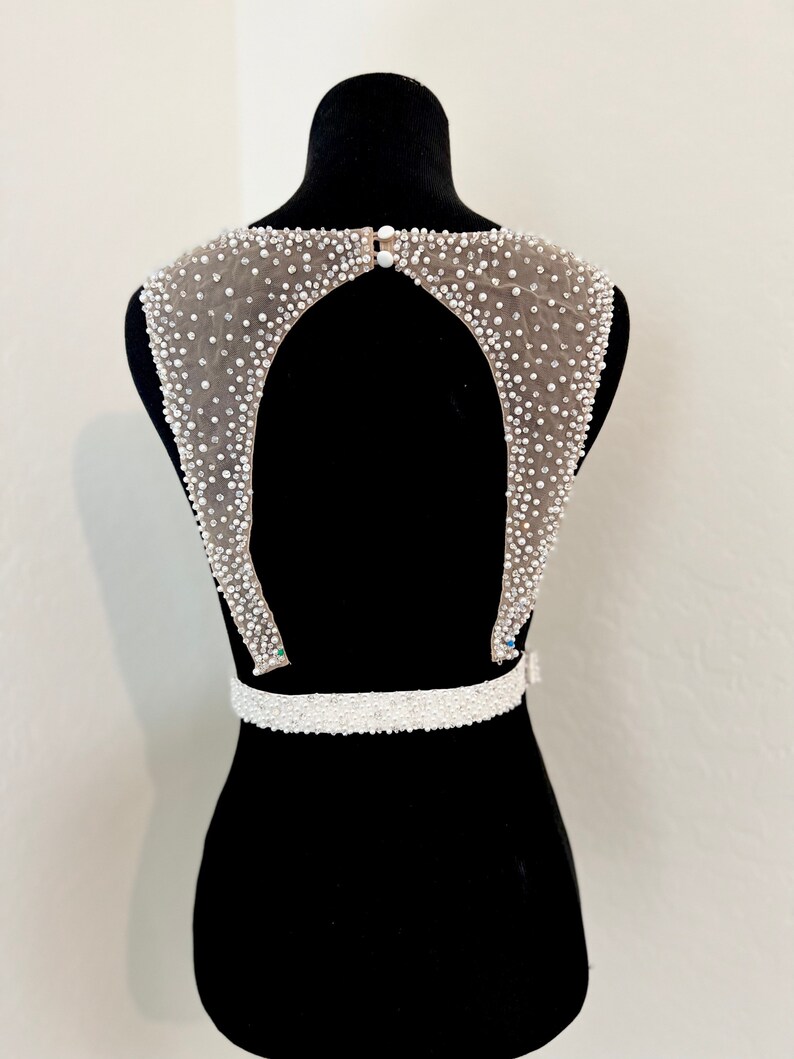 May include: A white beaded bridal belt with a low back design. The belt features a beaded neckline and a beaded waistband.