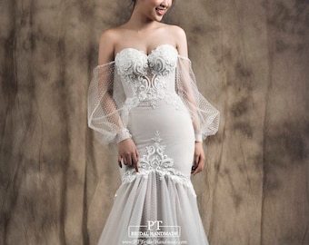 May include: A white off-the-shoulder wedding dress with a fitted bodice and a flowing skirt. The dress features intricate lace detailing and a sweetheart neckline. The sleeves are long and puffy, made of sheer fabric with polka dots. The dress is photographed against a textured brown background. PT Bridal Handmade www.PTBridalHandmade.com