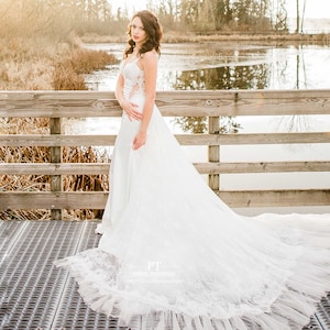 May include: A bride wearing a white lace wedding dress with a long train, standing on a wooden bridge over a pond. The bride is looking at the camera and smiling. The dress has a sweetheart neckline and a fitted bodice. The train is flowing behind her and is made of layers of tulle. The bride is wearing her hair in loose waves and has a natural makeup look. The background is a beautiful natural setting with trees and water.