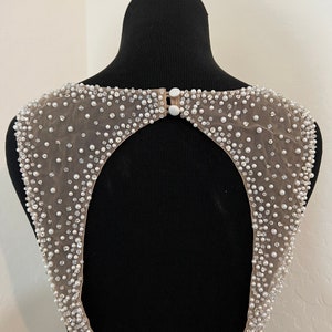 May include: A back view of a beaded, sheer fabric garment with a keyhole opening. The fabric is a light beige colour and the beads are white.