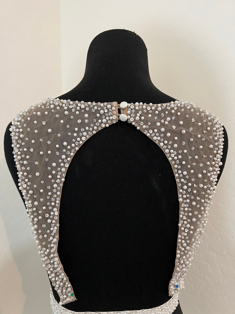 May include: A back view of a beaded, sheer fabric garment with a keyhole opening. The fabric is a light beige colour and the beads are white.