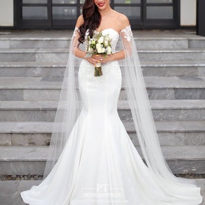 May include: A white off-the-shoulder mermaid wedding dress featuring a sweetheart neckline, intricate lace detailing on the bodice, and long, sheer tulle cape sleeves. The gown has a fitted silhouette that flares at the bottom into a train.