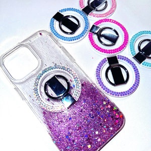May include: A clear phone case with a sparkly purple glitter design and a silver ring holder. The ring holder is surrounded by clear rhinestones. The case is shown with five additional ring holders in different colors: pink, blue, purple, and turquoise.