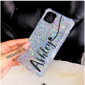 Pu&ograve; includere: Una custodia per telefono trasparente con uno sfondo di glitter argentati e il nome "Ashley" scritto in corsivo nero con un piccolo cuore sopra il nome.