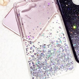 May include: A clear phone case with a glittery, iridescent design. The case has a pink and white background with silver glitter and stars. The text "iPhone" is printed on the case.