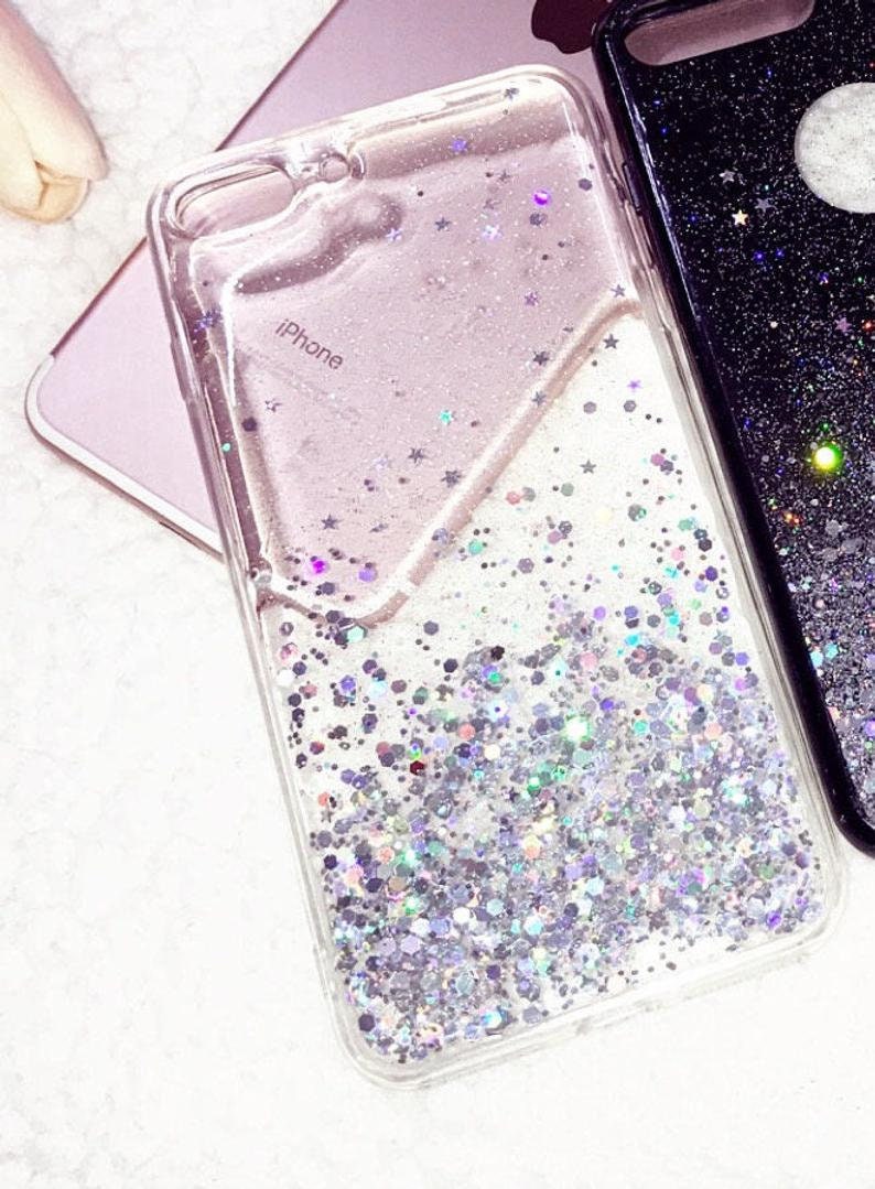 May include: A clear phone case with a glittery, iridescent design. The case has a pink and white background with silver glitter and stars. The text "iPhone" is printed on the case.