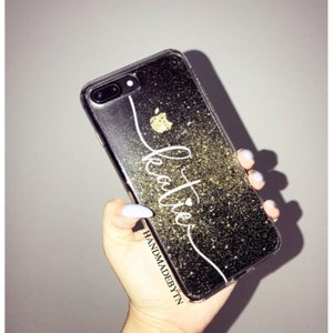 May include: A clear phone case with a black glitter background and a white cursive name "Katie" with a small heart. The case has a gold apple logo and the text "HANDMADEBYTTN" at the bottom.