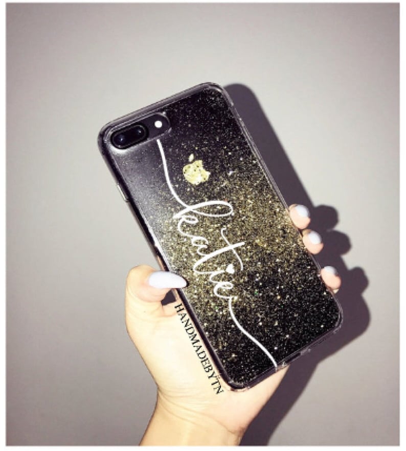 May include: A clear phone case with a black glitter background and a white cursive name "Katie" with a small heart. The case has a gold apple logo and the text "HANDMADEBYTTN" at the bottom.
