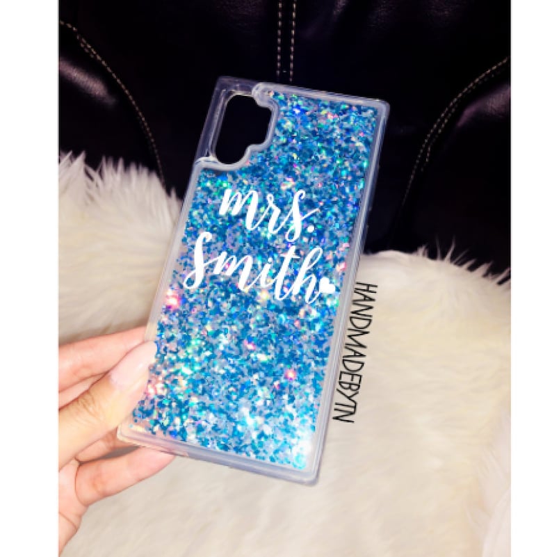 Pu&ograve; includere: Una custodia per telefono trasparente con un design a glitter blu e il testo "mrs. Smith" in bianco. La custodia &egrave; etichettata "HANDMADEBYTN".