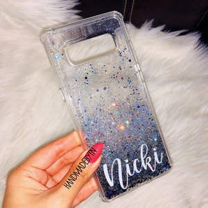 May include: A clear phone case with a silver glitter gradient and black glitter at the bottom. The case has the name "Nicki" written in white cursive lettering. The case is being held in a hand with red nail polish. The text "HANDMADEBYTN" is written on the hand.