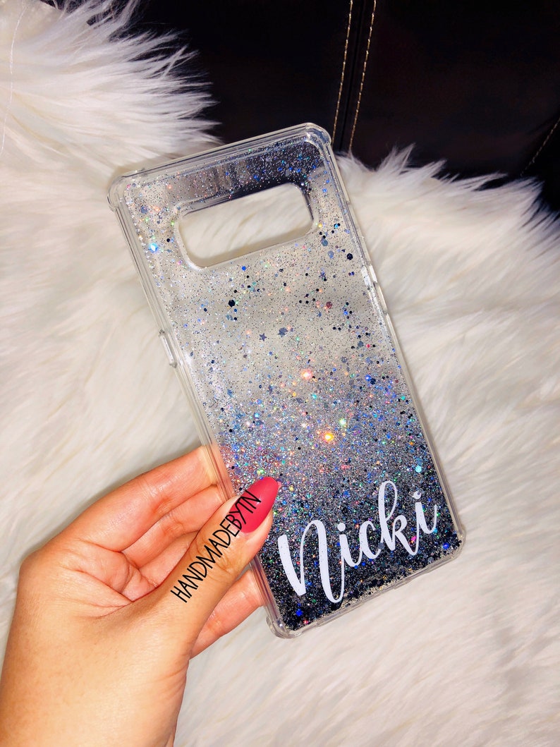 May include: A clear phone case with a silver glitter gradient and black glitter at the bottom. The case has the name "Nicki" written in white cursive lettering. The case is being held in a hand with red nail polish. The text "HANDMADEBYTN" is written on the hand.