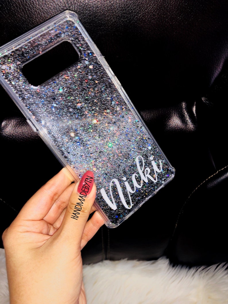 May include: A clear phone case with a silver glitter design and the name "Nicki" printed on the back. The case is being held by a hand with red nail polish.