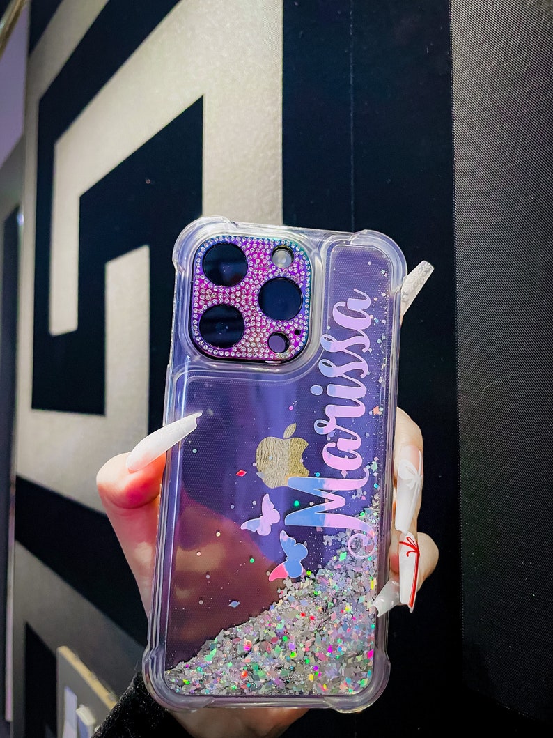 May include: A clear phone case with a glittery background and a purple, pink, and blue gradient. The case has a cutout for the camera lens, which is decorated with rhinestones. The case also has a gold apple logo and the name "Melissa" written on it.