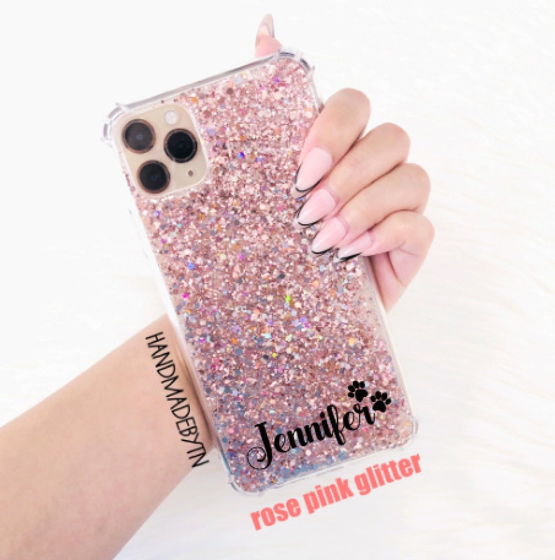 Pu&ograve; includere: Una custodia per telefono trasparente con un design glitter rosa. La custodia ha un'iscrizione nera che recita "Jennifer" con un simbolo di impronta di zampa. I testi "HANDMADEDBYTN" e "rose pink glitter" sono anche visibili.
