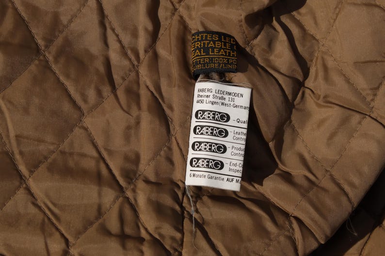 May include: Close-up of a brown quilted fabric with a tag attached. The tag has text that reads "ECHTES LEDER, ERITABLE REAL LEATHER". The tag also has the brand name "RABERG" and other text.