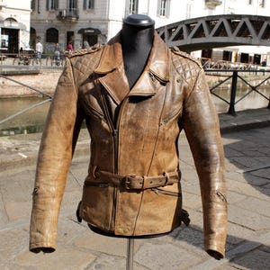 May include: A brown leather jacket with a belted waist and quilted shoulder details. The jacket has a zipper closure, multiple pockets, and a classic biker style. The jacket is displayed on a mannequin.
