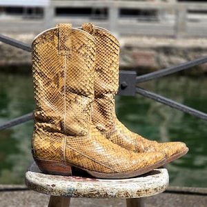 Op de afbeelding: Een paar hoge cowboylaarzen met een gestructureerd, goudbruin slangenleerpatroon. De laarzen hebben een puntige neus, een gestapelde houten hak en verticale stiksels langs de schacht. Het materiaal vertoont een duidelijk, herhalend schubbenpatroon.