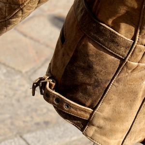 May include: Close-up of a brown leather jacket sleeve. The jacket features a quilted shoulder detail and a buckled strap at the cuff. The leather has a worn, textured appearance, with visible stitching and a weathered look.