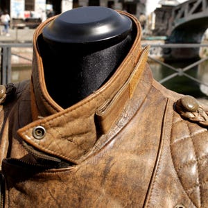 May include: A brown leather jacket with a quilted pattern on the shoulders and chest. The jacket has a notched collar, a zipper closure, and decorative braided shoulder accents. The jacket is displayed on a black mannequin head.