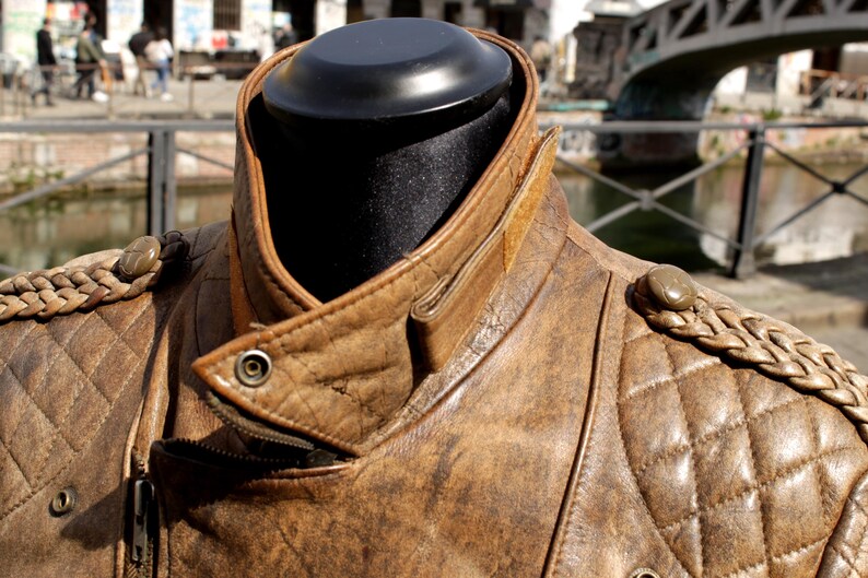 May include: A brown leather jacket with a quilted pattern on the shoulders and chest. The jacket has a notched collar, a zipper closure, and decorative braided shoulder accents. The jacket is displayed on a black mannequin head.