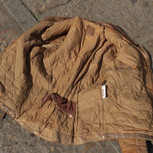 May include: A worn, brown leather jacket with a quilted, tan lining. The jacket is open, revealing the interior with pockets and a label. The leather shows signs of wear, adding to its vintage appeal. The jacket is lying on a stone surface.