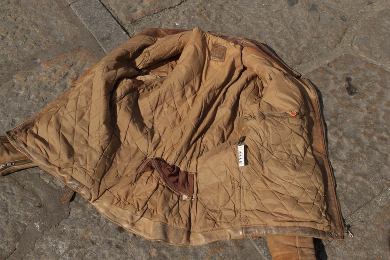 May include: A worn, brown leather jacket with a quilted, tan lining. The jacket is open, revealing the interior with pockets and a label. The leather shows signs of wear, adding to its vintage appeal. The jacket is lying on a stone surface.
