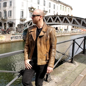 May include: A brown leather jacket with a quilted shoulder design. The jacket has a classic biker style with a notched collar and multiple pockets. The person is wearing black pants and sunglasses, standing near a canal with a bridge in the background.