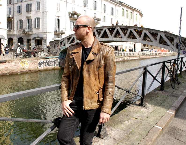 May include: A brown leather jacket with a quilted shoulder design. The jacket has a classic biker style with a notched collar and multiple pockets. The person is wearing black pants and sunglasses, standing near a canal with a bridge in the background.