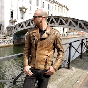 May include: A brown leather jacket with a quilted shoulder design and multiple pockets. The jacket has a zipper closure and a classic biker style. The person is wearing black jeans and sunglasses.