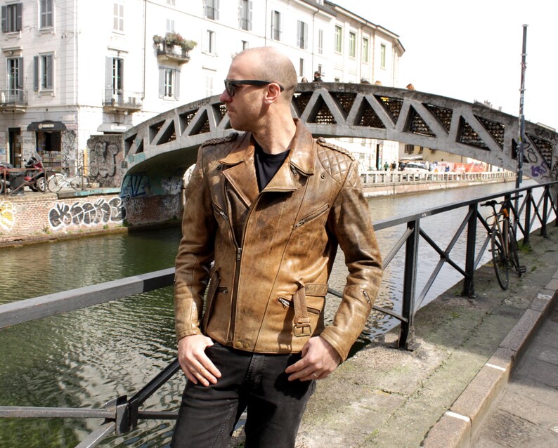May include: A brown leather jacket with a quilted shoulder design and multiple pockets. The jacket has a zipper closure and a classic biker style. The person is wearing black jeans and sunglasses.
