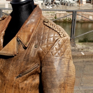 May include: A brown leather jacket with a distressed finish. The jacket features a quilted shoulder design, a notched collar, and multiple zippered pockets. The jacket is displayed on a black mannequin.
