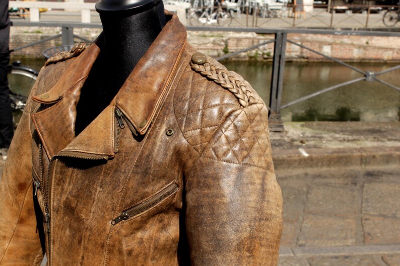 May include: A brown leather jacket with a distressed finish. The jacket features a quilted shoulder design, a notched collar, and multiple zippered pockets. The jacket is displayed on a black mannequin.
