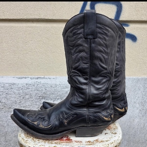 Peut inclure: Une paire de bottes de cow-boy en cuir noir avec des motifs de surpiqûres ton sur ton sur la tige haute et l'empeigne. Les bottes présentent un bout pointu, un talon bottier et de subtils accents dorés intégrés aux coutures décoratives.