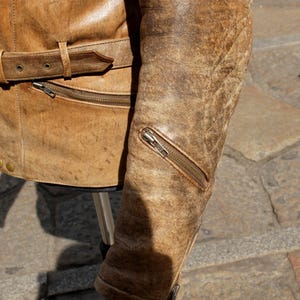 May include: Close-up of a distressed brown leather jacket. The jacket features a belt at the waist, a zippered pocket, and a zipper on the sleeve. The leather has a worn, textured appearance.