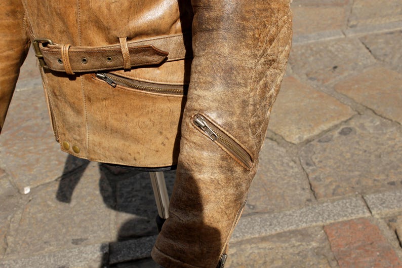 May include: Close-up of a distressed brown leather jacket. The jacket features a belt at the waist, a zippered pocket, and a zipper on the sleeve. The leather has a worn, textured appearance.