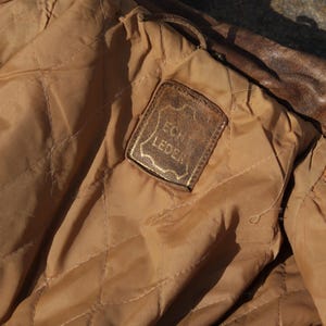 May include: Close-up of the interior of a brown leather jacket. The lining is a quilted tan fabric. A rectangular leather tag with the words "Echt Leder" in gold lettering is sewn into the lining.