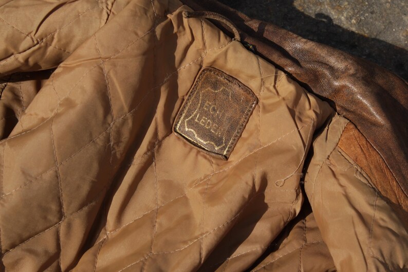 May include: Close-up of the interior of a brown leather jacket. The lining is a quilted tan fabric. A rectangular leather tag with the words "Echt Leder" in gold lettering is sewn into the lining.