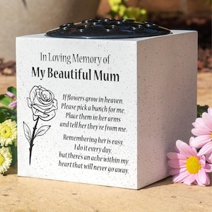 May include: A white memorial cube with a black floral arrangement holder. The cube is inscribed with "In Loving Memory of My Beautiful Mum" and a poem, alongside a rose illustration. Pink and yellow flowers are in the background.