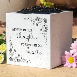 May include: White square memorial vase with a black plastic insert for flowers. The vase has a floral design and the text "Always in our thoughts forever in our hearts".
