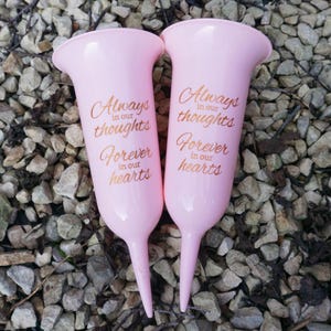 May include: Two pink plastic flower vases with a pointed base. The vases have the text "Always in our thoughts Forever in our hearts" printed on them in gold.