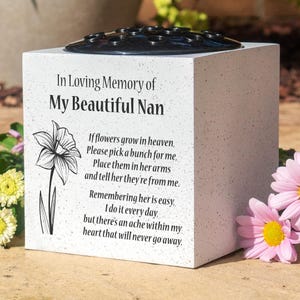 May include: A white memorial flower vase with a black floral grid top. The vase has the text "In Loving Memory of My Beautiful Nan" and a poem with a daffodil illustration. Pink daisies are in the foreground.