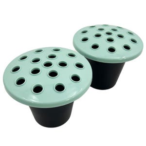 May include: Two flower frogs, each with a mint green top and a black base. The tops are round with multiple holes for arranging flowers. The bases are cone-shaped. The frogs are designed to hold flowers in place.