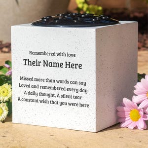 May include: A square, white memorial vase with a black floral grid on top. The vase is inscribed with the words "Remembered with love" and "Their Name Here". Further text includes "Missed more than words can say". Pink and yellow flowers are visible.