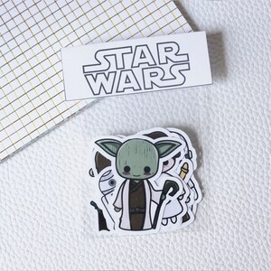 Puede incluir: Una bolsa de plástico transparente con una etiqueta blanca que dice "STAR WARS" y contiene un conjunto de cinco pegatinas de personajes de dibujos animados. Las pegatinas representan a Yoda, R2-D2, un Stormtrooper, la princesa Leia y Darth Vader.