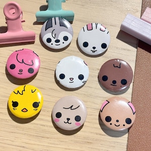 Puede incluir: Una colección de nueve botones redondos y coloridos con caras de animales de dibujos animados. Los botones son de varios colores, incluyendo rosa, amarillo, marrón y blanco. También se incluyen dos clips de encuadernación y dos pequeños clips de madera.