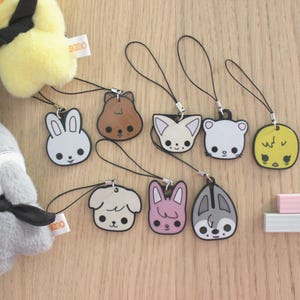 May include: A collection of cartoon animal face charms with black outlines and attached black cords. The charms include a white bunny, brown bear, white fox, white bear, yellow chick, white dog, pink bunny, and gray bunny. A yellow plush chick and gray plush animal are also visible.