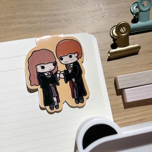 Puede incluir: Una pegatina colorida con personajes de dibujos animados con túnicas negras tomados de la mano. La pegatina está colocada en la página de un cuaderno. Otros elementos incluyen un clip dorado, un clip turquesa y dos marcadores rectangulares.